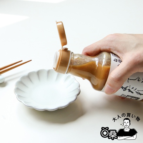 一只手拿著裝有芝麻沾醬的玻璃瓶，瓶蓋打開，醬料正從瓶中倒入一個白色、帶有花瓣狀邊緣的淺碟中，碟旁擺放著兩根木質筷子。
