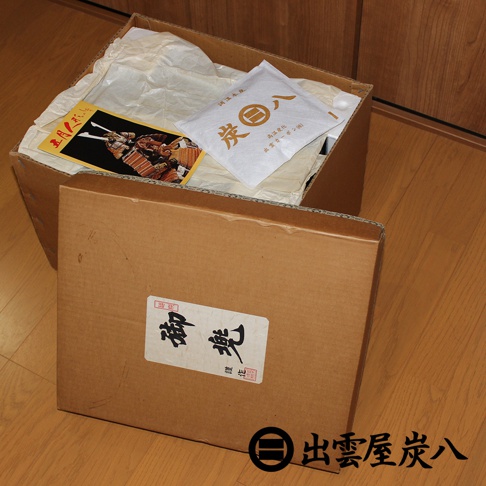 一個棕色紙箱放在木質地板上，箱子內部鋪著白色襯紙，放有一包印有出雲屋炭八字樣的調濕木炭包，以及一張印有武士盔甲圖案的精美卡片。前方斜靠著一個貼有御兜書法字樣標籤的紙板蓋，右下角則標示著出雲屋炭八的品牌名稱與標誌。