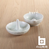 兩個白色陶瓷小碟，其中一個碟中央有直立的鳥形支架，另一個碟則有鳥嘴造型，兩者均有精緻的紋理裝飾，置於木質背景上。