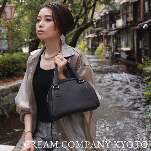 一位女性手持一個黑色皮革製成的半月形迷你手提包，包包上有白色車縫線，搭配淺色絲質外套和灰色褲子，背景是古樸的日式街景和流水。