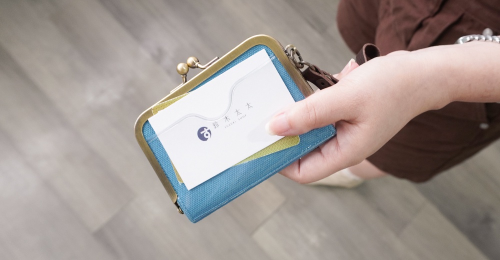 一個女性的手拿著一個藍色帆布製、帶有金色金屬扣的卡片包，裡面放著一張印有日文和英文「SUZUKI SHOP」的白色名片。