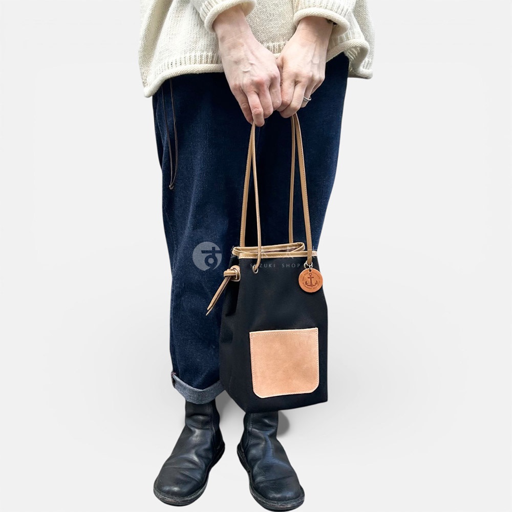 一個穿著白色毛衣和藍色牛仔褲的人，手裡拿著一個黑色束口包，包上有一個淺棕色的皮革口袋和一個吊牌。