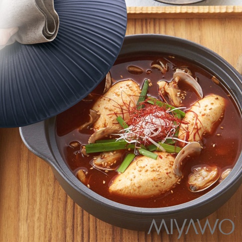 深灰色陶土湯鍋盛有紅湯燉菜，鍋內有豆腐、蛤蜊、蔥段和紅椒絲。鍋蓋為深灰色，帶有放射狀紋理，一角有淺色布料。