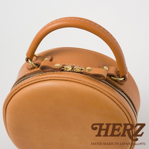 HERZ 品牌標誌與 Since 1973 字樣，搭配手工皮革圓形側背包的提把及拉鍊細節