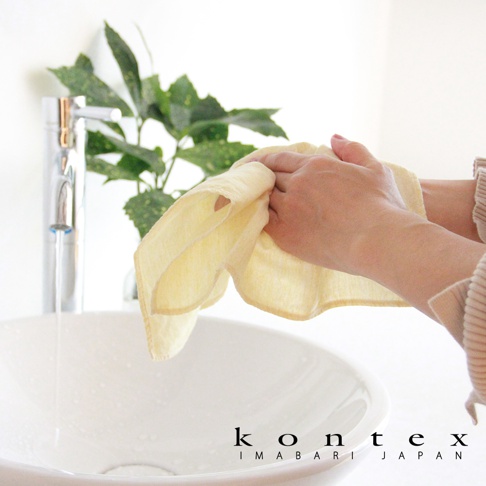 一塊淺黃色的 MOKU LINEN 棉麻拭巾，輕薄且具有吸水性，由雙手拿著，旁邊有水龍頭和洗手盆。