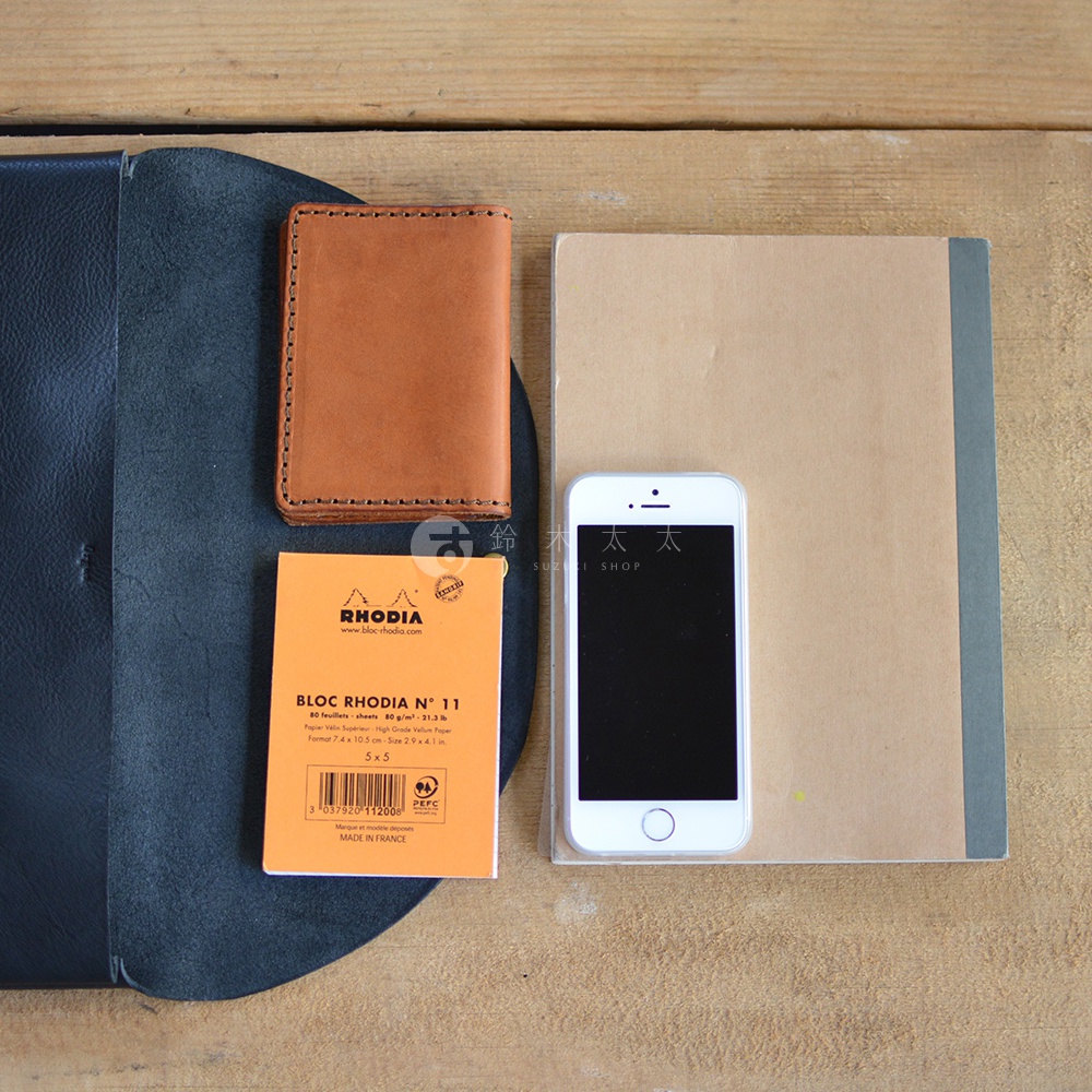 深藍色皮革內頁、棕色皮革小物收納袋、白色手機、橘色筆記本，擺放在木紋桌面上。