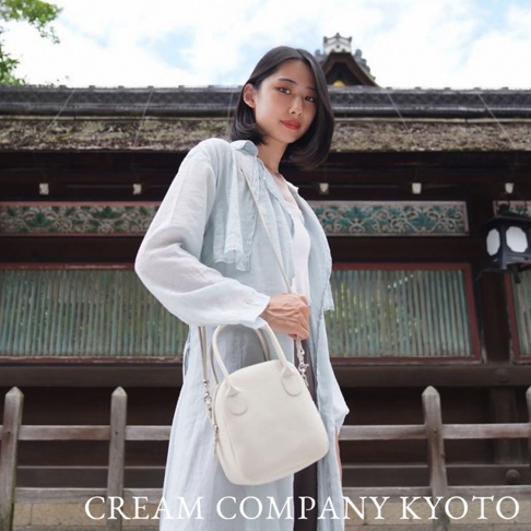 一位女子身穿淺藍色長袖上衣，肩背一個奶油色皮革貝殼包，包包上有手提帶和可拆卸的肩帶，整體呈現簡約風格。