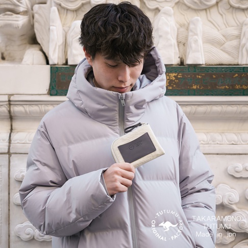 一位年輕男性身穿淺灰色連帽鋪棉外套，左手拿著一個米白色零錢包，包上印有TAKArAMONO TUTUMU Made in jpn的字樣。