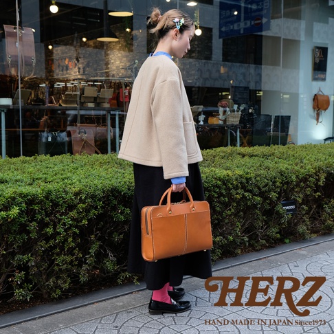 駝色皮革雙口袋兩用公事包，附有手提把與可拆卸肩帶，設計簡潔，適合商務場合。