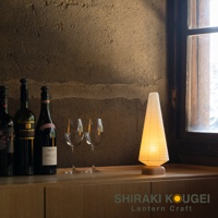 八女迷你裝飾提燈，錐形燈罩由白色紙質製成，帶有木質底座，散發溫暖的黃光。旁邊擺放有紅酒瓶與酒杯，背景為粗糙的土牆與木框窗戶。