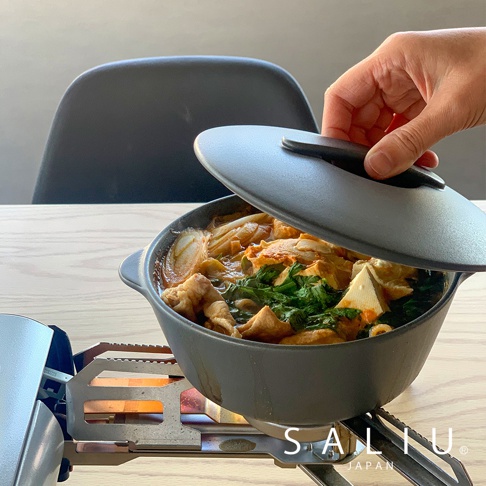 一個灰色的耐熱陶製圓鍋，鍋內裝有燉煮的食物，包括肉片、蔬菜和豆腐，鍋蓋被一隻手提起，露出鍋內食材。鍋子放在一個銀色的戶外爐具上，爐火正在燃燒。