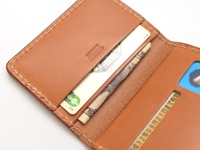 職人手作皮革雙折證件收納夾，採用棕褐色皮革製成，內部可見卡片、日圓紙鈔及印有日文的證件。車縫線明顯，整體呈現質感。