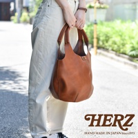 一位女士手持一個棕色皮革製的圓形肩掛托特包，包包上有縫線細節與金屬扣環，搭配白色長褲和棕色手錶。