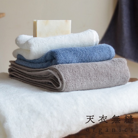 堆疊的 Lisse 蓬鬆柔感有機棉浴巾，包含白色、藍色和灰色。左上方有一塊淺色肥皂。