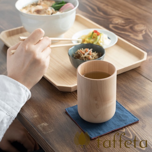 淺色楓木製水杯，杯身呈現自然木紋，盛有琥珀色茶飲，放置於深藍色杯墊上。