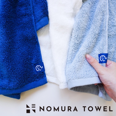野村毛巾 SUUZO 系列，展示深藍、白色及淺藍三色超無撚極吸水半浴巾，其中淺藍色浴巾一角有手部撫摸的細節，另有野村毛巾的品牌標誌。