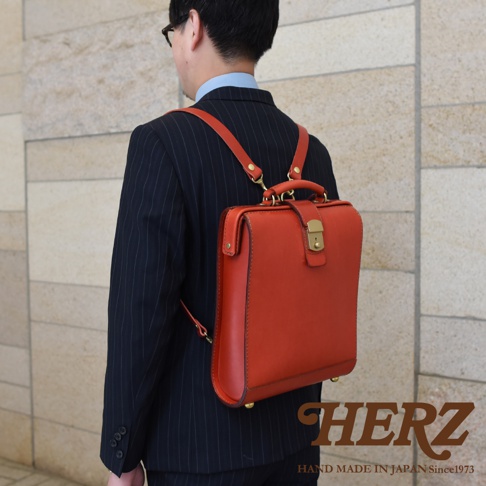 一名男性身穿深色條紋西裝，背著一個紅色皮革製成的杜勒斯商務後背包。此款後背包採用簡潔的設計，具有一個金屬鎖扣和頂部提把，整體呈現復古風格。