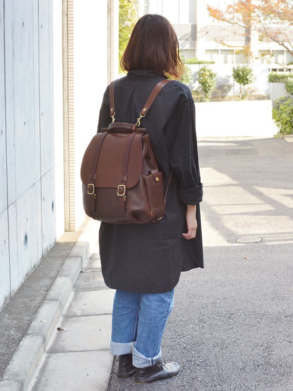 深棕色皮革雙扣後背包，設有頂部提把、兩個前袋和可調節肩帶，搭配黑色長版上衣和捲褲腳牛仔褲。