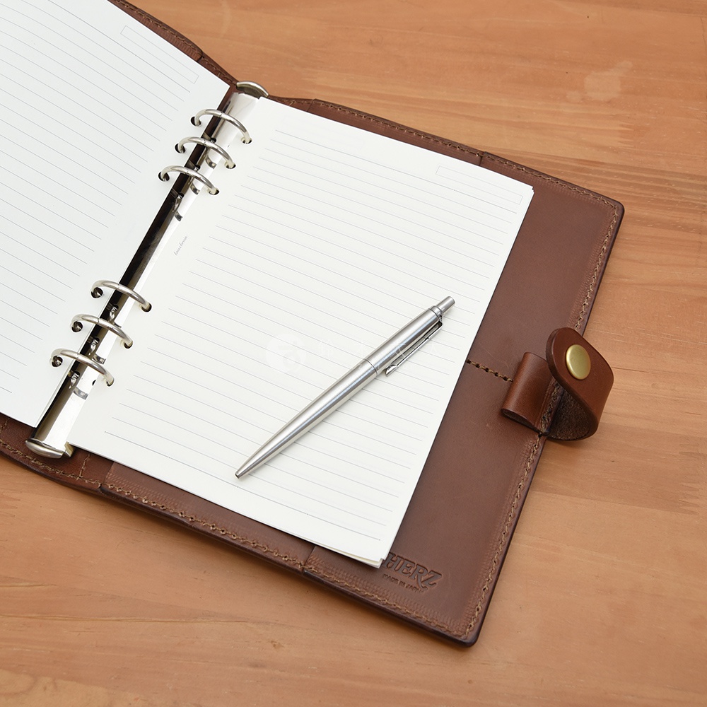 棕色皮革A5手帳/記事本書套，內有線條內頁及一支銀色金屬原子筆，書套右側有一皮革繫帶及黃銅色鈕扣。