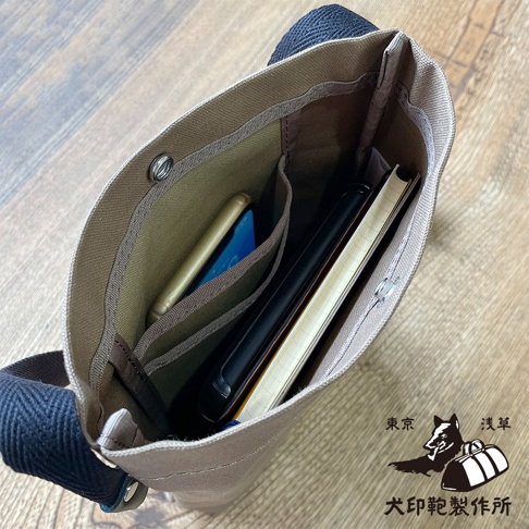 帆布材質的平板收納側背包內部，可見手機、筆記本等物品，背包側邊印有東京淺草犬印鞄製作所的標誌。