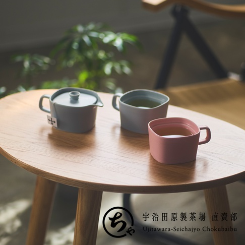 一張放在淺色木質圓桌上的生活場景照片，桌面擺放著宇治田原製茶場的簡約美濃燒茶具，包含一個灰色的個人用茶壺，以及兩個分別為灰色和粉色的方形馬克杯，後方帶有模糊的植栽綠意與椅子線條，整體呈現出溫馨舒適的飲茶氛圍。