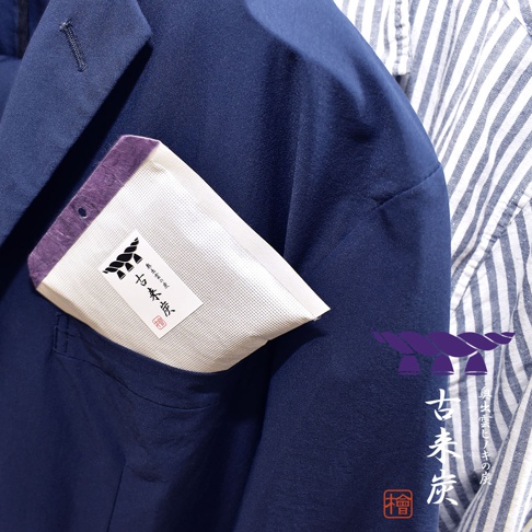 深藍色西裝外套的胸前口袋中，放置著一個白色網狀材質的調濕木炭包，頂部邊緣為紫色。木炭包上印有黑色繩結圖案及日文文字。