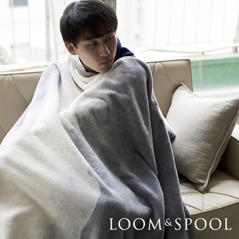 一位男子蓋著一件抗菌短絨單人毯，毯子為漸層灰白色，質地柔軟，蓋在米色沙發上，旁邊有一個淺灰色抱枕。