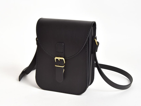 黑色皮革翻蓋迷你單肩包，配有可調節式肩帶和金色針扣。包身線條簡潔，手工縫線清晰可見。