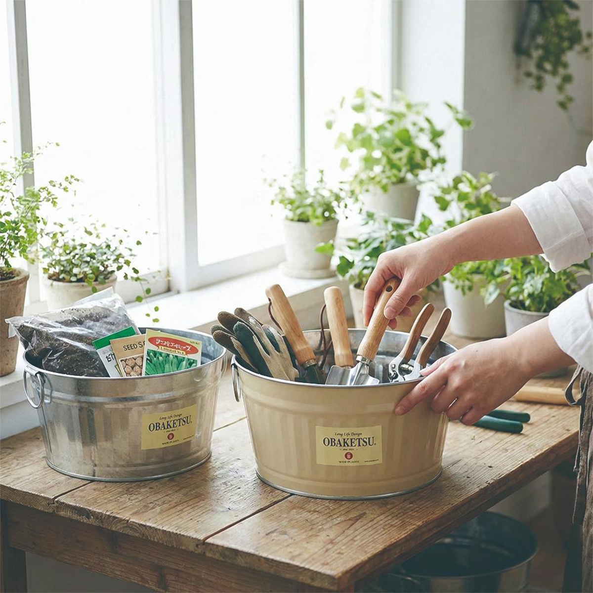 模式B： 兩個復古風格的OBAKETSU收納桶，擺放在木桌上，其中一個裝滿園藝工具，另一個則裝著土壤和種子包，後方窗台邊擺放著綠色盆栽。