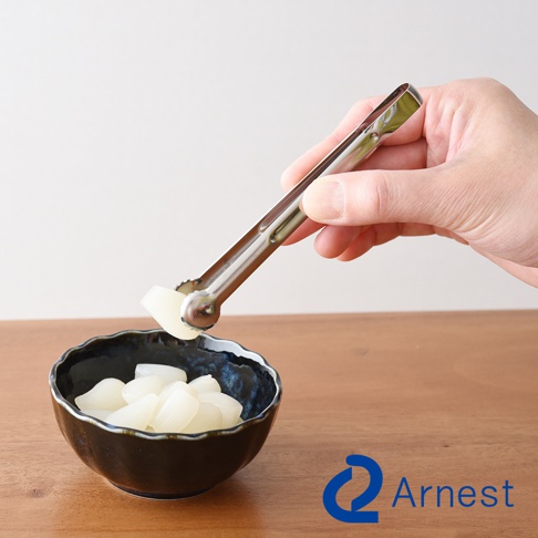 手持不鏽鋼萬用料理夾，夾取一塊白色食材，上方為白色背景，下方為木質桌面及一個黑色紋理小碗，碗內盛有白色塊狀食材。