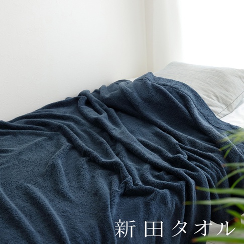 一張展示新田毛巾品牌泉州無漿毛巾單人被的床鋪特寫，柔軟深藍色的毛巾被覆蓋在床墊上，呈現出自然的褶皺感與溫暖質感，後方搭配一顆灰色的枕頭，畫面右下角附有新田タオル字樣。