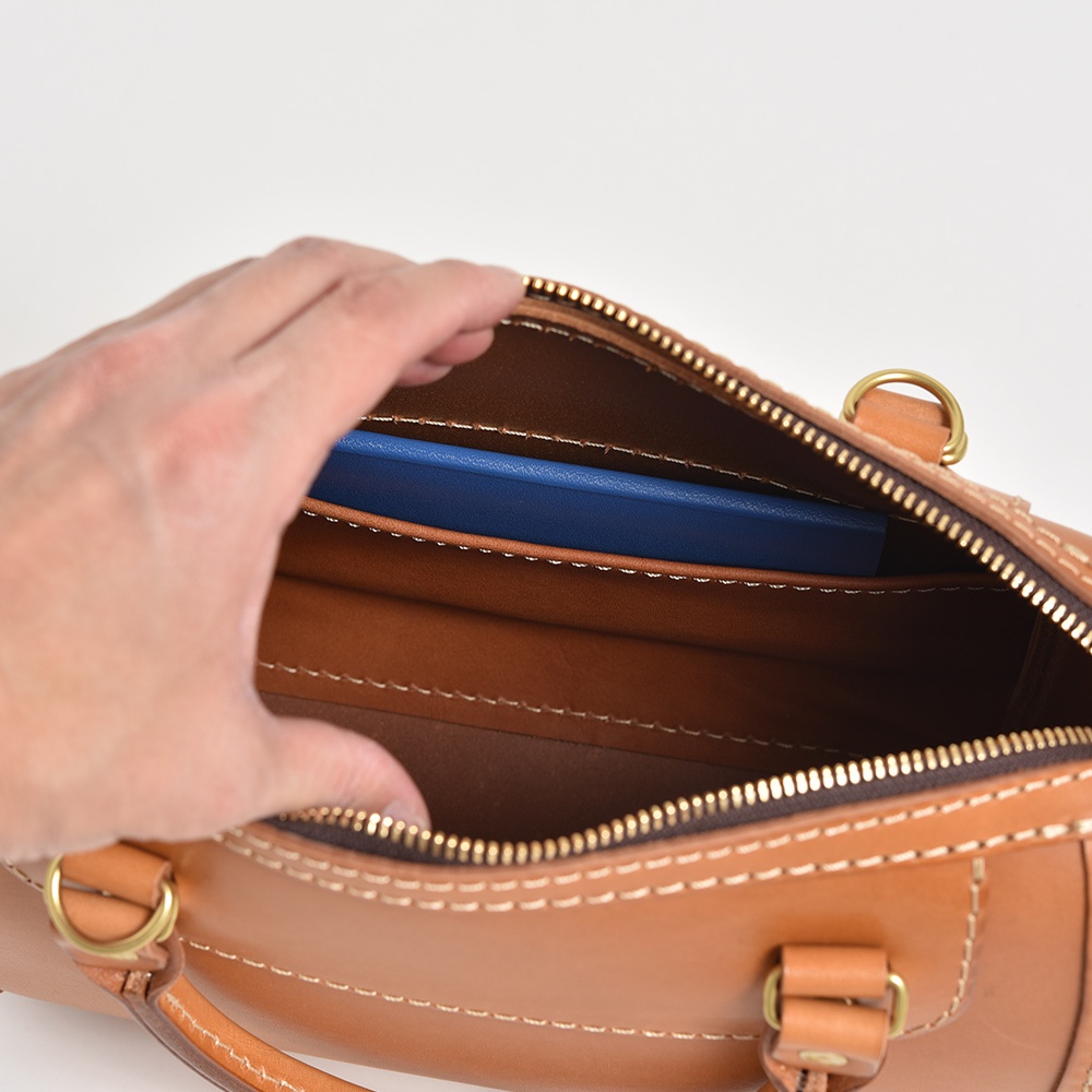 職人手作皮革迷你波士頓包，採用天然皮革材質，呈現溫潤的棕褐色。包款開口處有金色拉鍊，內層可見藍色長夾，包體外部縫線明顯，結構穩固。