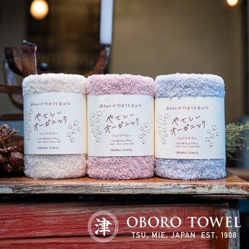 三條捲起的有機棉毛巾，依序為米白色、淺粉色與淺藍色，毛巾質地蓬鬆柔軟，包裝紙帶上有日文說明與棉花圖案。