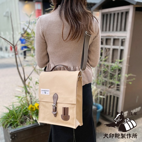 一位女士身揹一個米色純綿帆布製成的直式側背包，包包上有棕色皮革提把和束帶，側邊有兩個小皮製插槽。