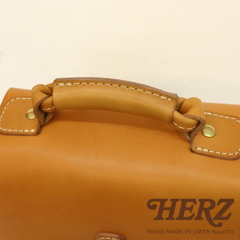 近距離特寫職人手作皮革單肩兩用小方包，展現溫潤的天然皮革紋理與精緻車縫細節。包款為溫暖的淺棕色調，配有堅固的皮革提把，提把兩端以同色皮革繩結固定，並有金屬卯釘裝飾。