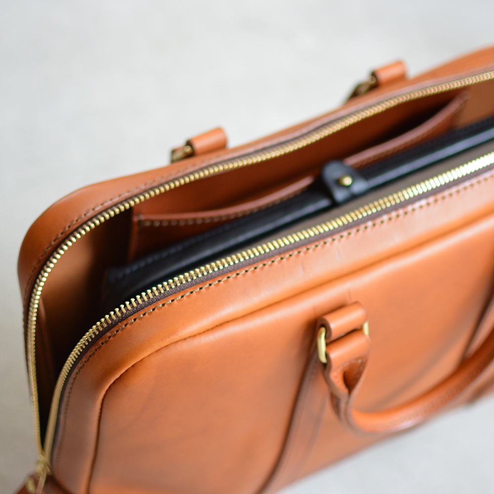 棕色皮革公事包，拉鍊部分打開，內部可見黑色文件夾或筆記本，配有金色拉鍊和金屬扣飾，以及兩個短提把。