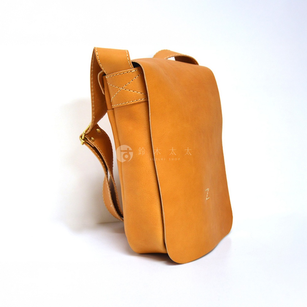 一個由黃褐色皮革製成的斜背包，有著一個翻蓋和一個可調節的肩帶，背包側面印有微小的標誌。