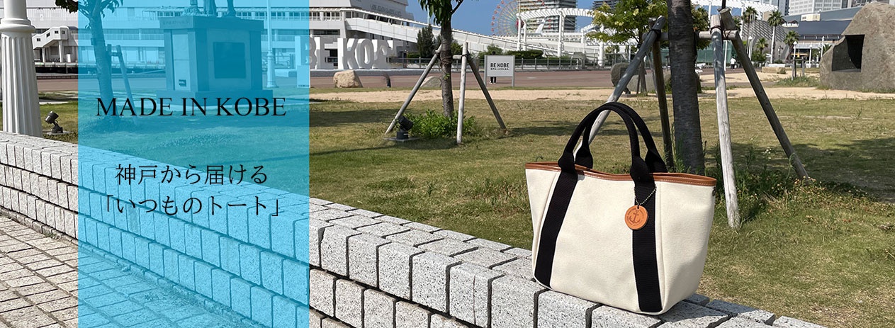 米白色帆布手提托特包，配有黑色織帶和棕色皮革飾邊。包側有兩個黑色織帶裝飾，包上掛有圓形皮革吊飾，上面印有船錨圖案。