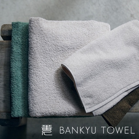 三條不同顏色（綠色、淺灰色、深棕色）的柔軟毛巾疊放在木質表面上，毛巾質地蓬鬆，顯示出其吸水性。