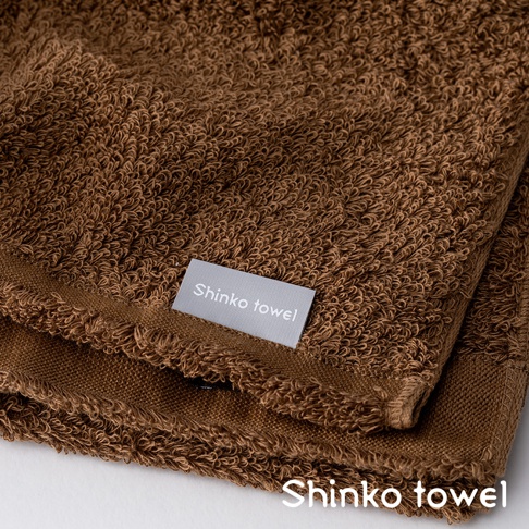 MAISON BLANC 輕奢柔膚匹馬棉毛巾，呈現溫暖的棕色調，毛圈蓬鬆細緻，觸感柔軟。毛巾一角縫有淺灰色標籤，上面印有「Shinko towel」字樣。