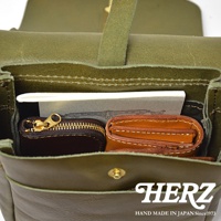 HERZ 品牌標誌與日本製造 since 1973 字樣，置於綠色皮革包的下方。包內可見一個棕色零錢包與一個棕色皮夾。