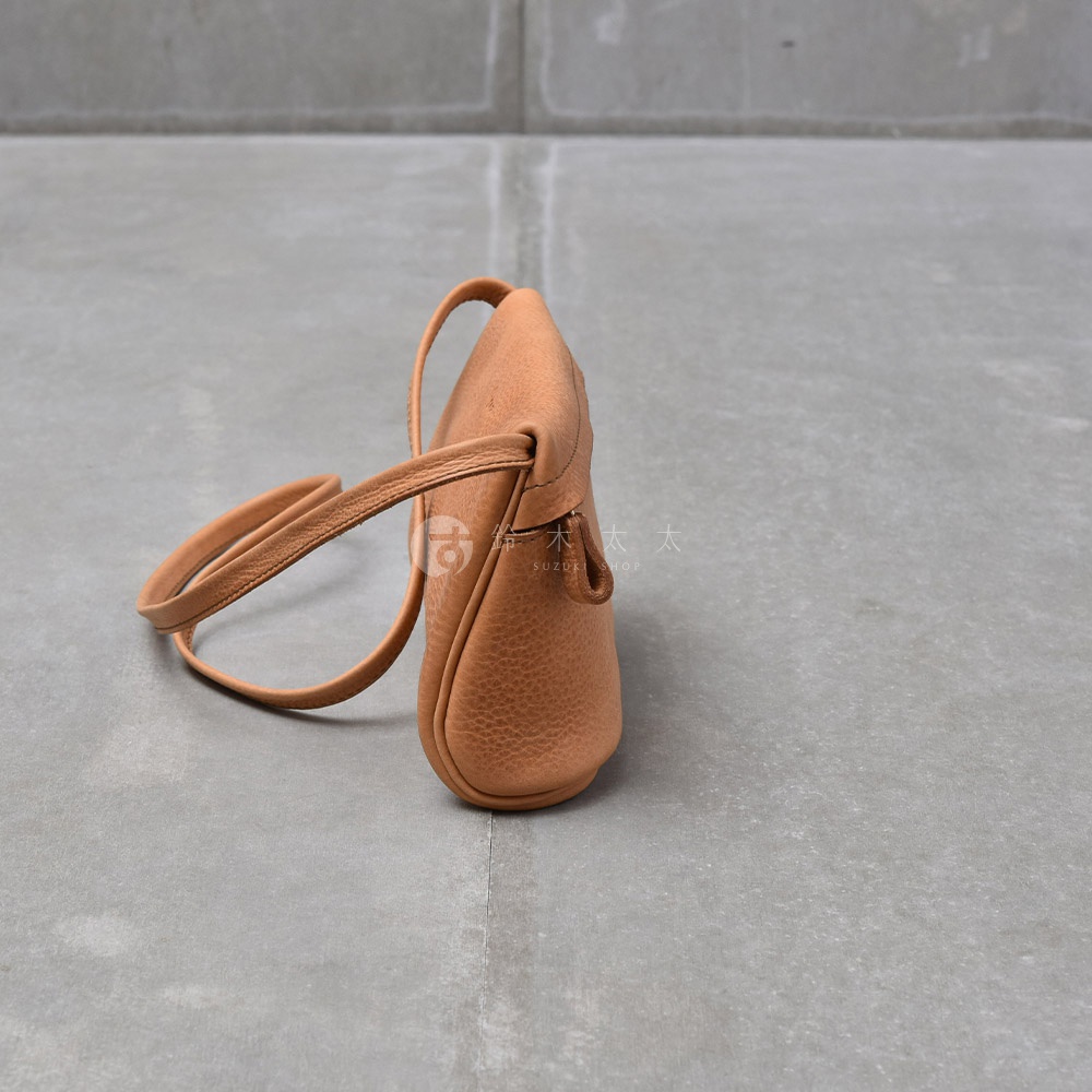 一個淺褐色的皮革斜背包，有著自然紋理，包身呈現圓潤的曲線，搭配細長的同色系背帶，置於灰色水泥地面上。