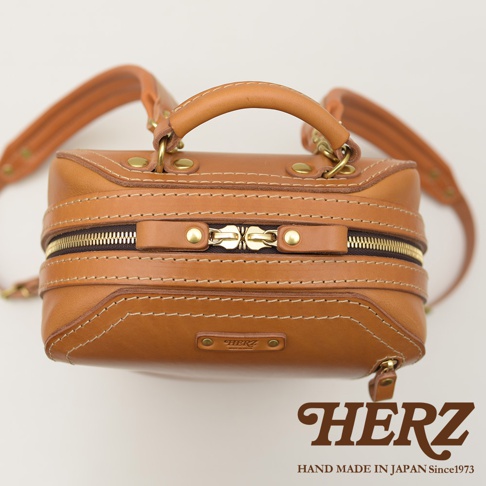 一個棕褐色的皮革立體商務後背包，配有雙向拉鍊、兩個提把和可調節肩帶。包身上有多處縫線和金屬鉚釘裝飾，左下方有一個標有「HERZ」的皮革標籤。