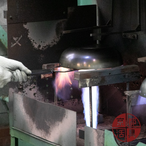 一個戴著白色手套的手，正操作著一個平底鐵鍋，鍋身呈現金屬光澤。鍋底下方有火焰，火焰顏色從橙紅色漸變到藍色，顯示正在加熱的過程。