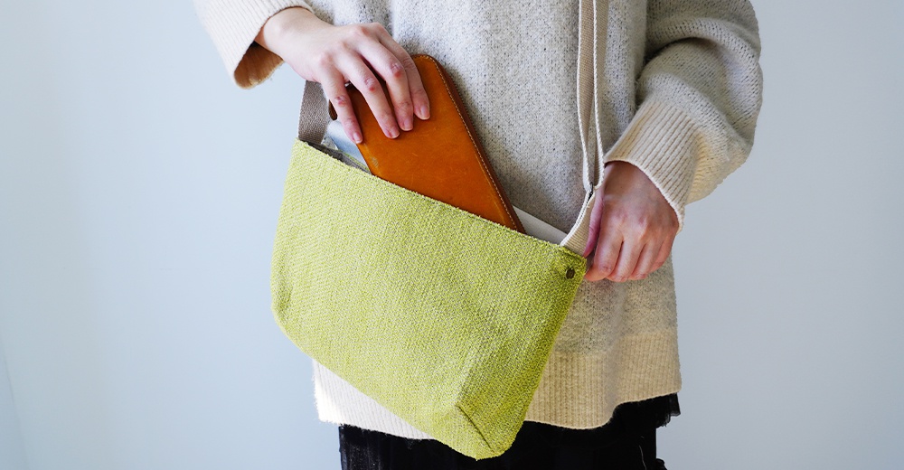 穿著米白色毛衣的人，手將一個橘色長夾放入淺綠色紋理手提袋。