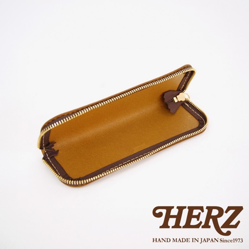 HERZ 品牌標誌，下方為 HAND MADE IN JAPAN Since1973 字樣。一個打開的棕色皮革筆袋，拉鍊為金色。