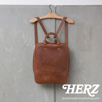 一個棕色的皮革後背包，掛在木製衣架上，背景是灰色牆壁，右下角有HERZ品牌標誌。