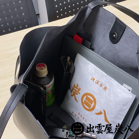 黑色皮革手提包內部，放置了一瓶綠色包裝的飲品、一個黑色皮夾，以及一包印有出雲屋炭八字樣的白色方形調濕木炭，包底還有鑰匙圈與鑰匙。