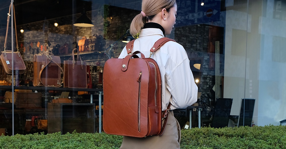 棕色皮革手作後背包，方形設計，正面有一個拉鍊口袋和一個旋鈕扣，肩帶可調節，適合商務休閒風格。