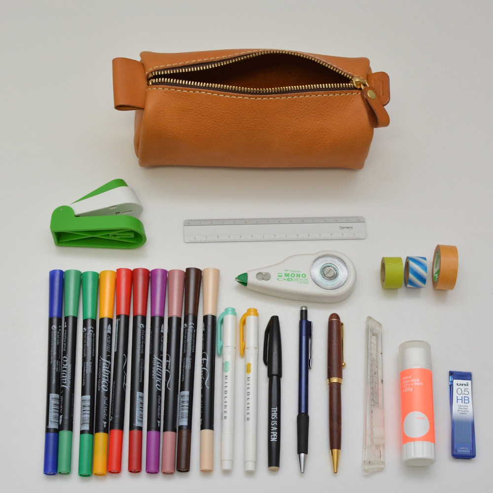 職人手作皮革多用途收納筆袋，顏色為棕色，搭配金色拉鍊。周圍擺放有綠色小釘書機、尺、修正帶、綠色、藍白色條紋、橘色膠帶、共13支彩色馬克筆、兩支白色螢光筆、一支黑色簽字筆、一支藍色簽字筆、一支棕色鋼筆、一支美工刀、一支橘色膠水和一支藍色筆芯。